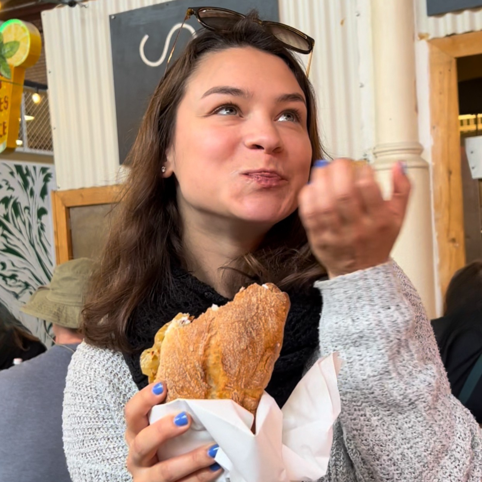 Lena isst ein belegtes Brot in einem Café und lächelt. Food-Content und Genussmoment in natürlicher Alltagsszene.  Lena eating a sandwich in a café and smiling. Food content and enjoyment in a natural everyday moment.